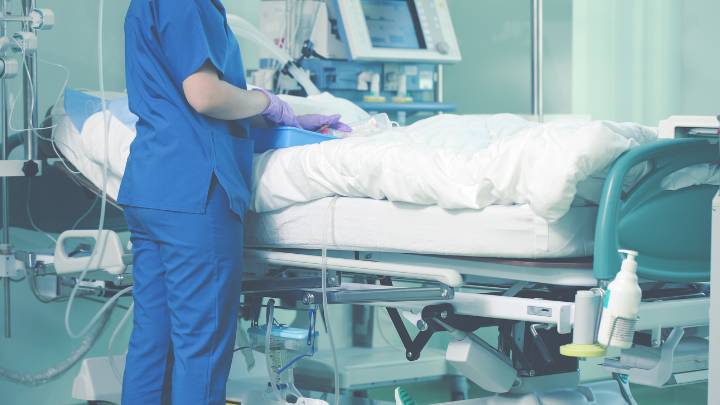 A woman in blue scrubs stands next to a hospital bed.